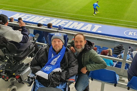 Zwei BUDDIES mit und ohne Rollstuhl lächeln im Stadion des KSC in die Kamera. Im hintergrund sieht man das Spielfeld