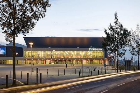 Im Halbdunkel leuchtet die Aussenfassade der modernen EWE Arena Oldenburg. Die Straße davor, ist komplett leer.