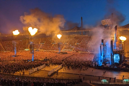 Ein breites Publikum steht vor einer Bühne im Stadion. In der Luft schweben flammende Herzen