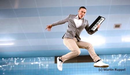 Andreas Langsch springt in die Luft vor einem blauen Hintergrund. In der linken Hand hält er ein Mini-Keyboard