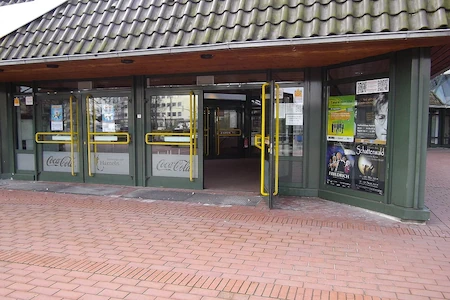 Der Eingangsbereich der rattenfängerhalle Hamlen ist ebenerdig. Der Zugangsweg ist mit roten Backsteinen ausgelegt. Eine der breiten Glastüren steht offen. Fenster und Türen sind mit Plakaten für kommende Veranstaltungenn behängt.