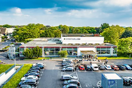 Ein Blick auf die Stadthalle Köln. Es ist der Ausbereich und die Fassade zu sehen, davor ein Parkplatz mit einigen Autos.