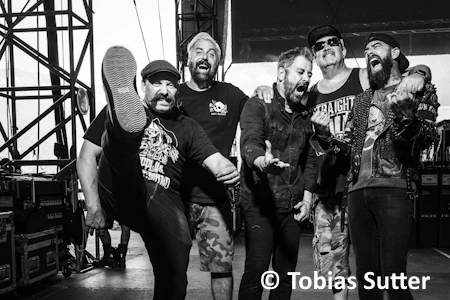 Die Band Zebrahead hinter der Bühne. Das Bild ist in Schwar Weiß gehalten. Alle haben Spaß und die unterschiedlichsten Gesichtsausdrücke. Einer hat das Beinn in in Richtung Kamera wie bei einem Kick in die Luft gestreckt.