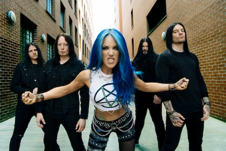 Die Sängerin von Arch Enemy hat blaue Haare. Sie zieht ein böses Gesicht und hat die Arme bedrohlich ausgebreitet. Hinter ihr steht mit einigem Abstand die Band