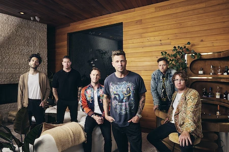 Die Bandmitglieder von OneRepublic sitzen gemütlich herum. Sie schauen in die Kamera, nur der Sänger steht.