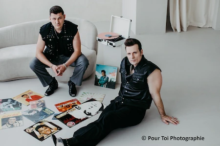 Die zwei Musiker von The Baseballs im stilechten 50 Jahre Outfit mit den passenden Frisuren. Sie sitzen in einem hellem Raum, einer auf einem Sofa einer auf dem Boden. Ein altmodischer Schallplattenspieler mit Platten von Elvis ist ebenfalls zu sehen.