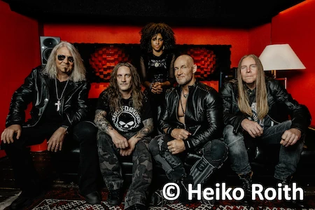 Die deutsche Heavy und Power Metal Band Primal Fear sitzt auf einem Sofa, die neue Gitarristin steht im Hintergrund