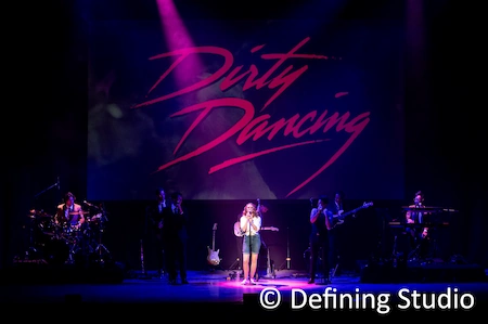 Auf der großen Bühne sieht man das Logo von Dirty Dancing in Concert, davor eine Sängerin und einige Musiker im Rampenlicht